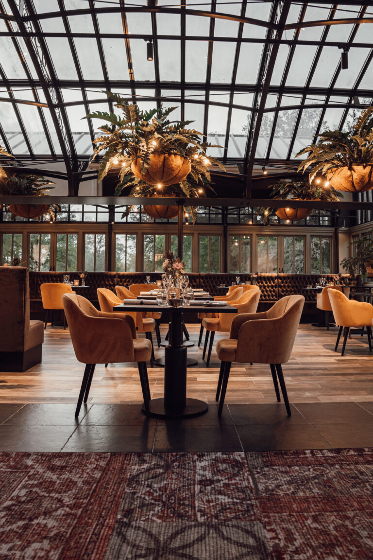 Sfeervolle restaurantserre met glazen dak, gedekte tafels, oranje fluwelen stoelen en verlichte hangplanten.