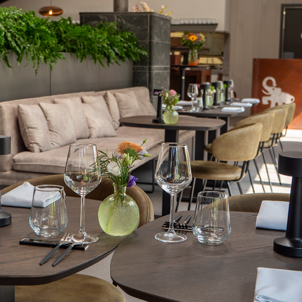 Modern restaurant interieur met elegant gedekte tafels, zwarte tafellampen, beige banken en groene planten.