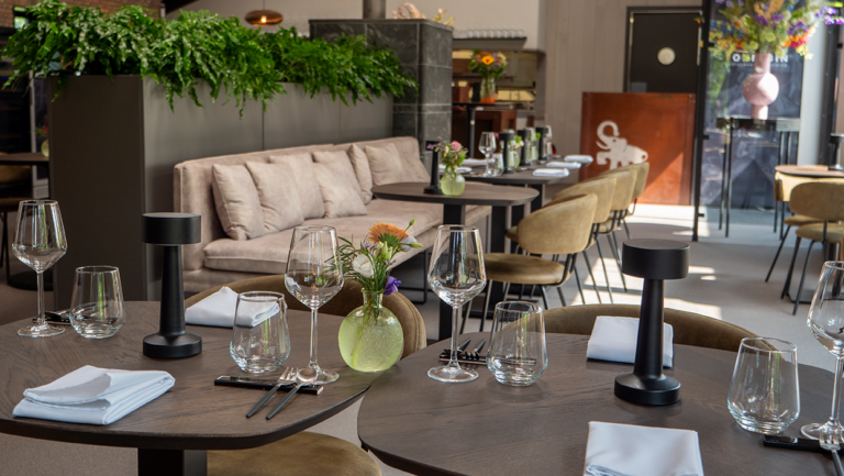 Modern restaurant interieur met elegant gedekte tafels, zwarte tafellampen, beige banken en groene planten.