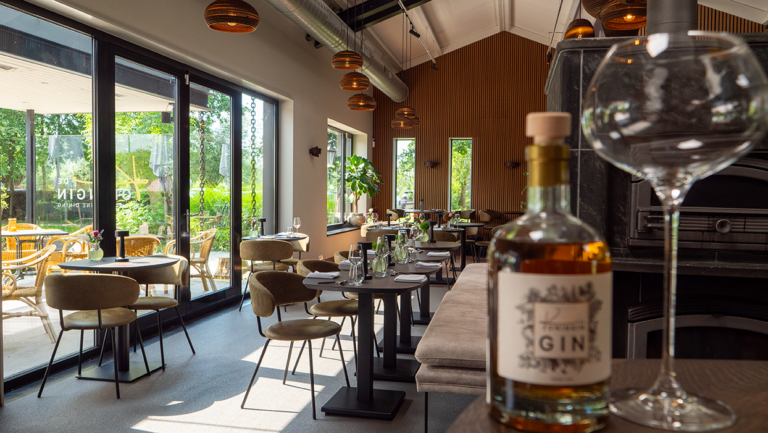 Fles GIN en glas op houten tafel in een modern restaurant met houten muren en uitzicht op groen terras.