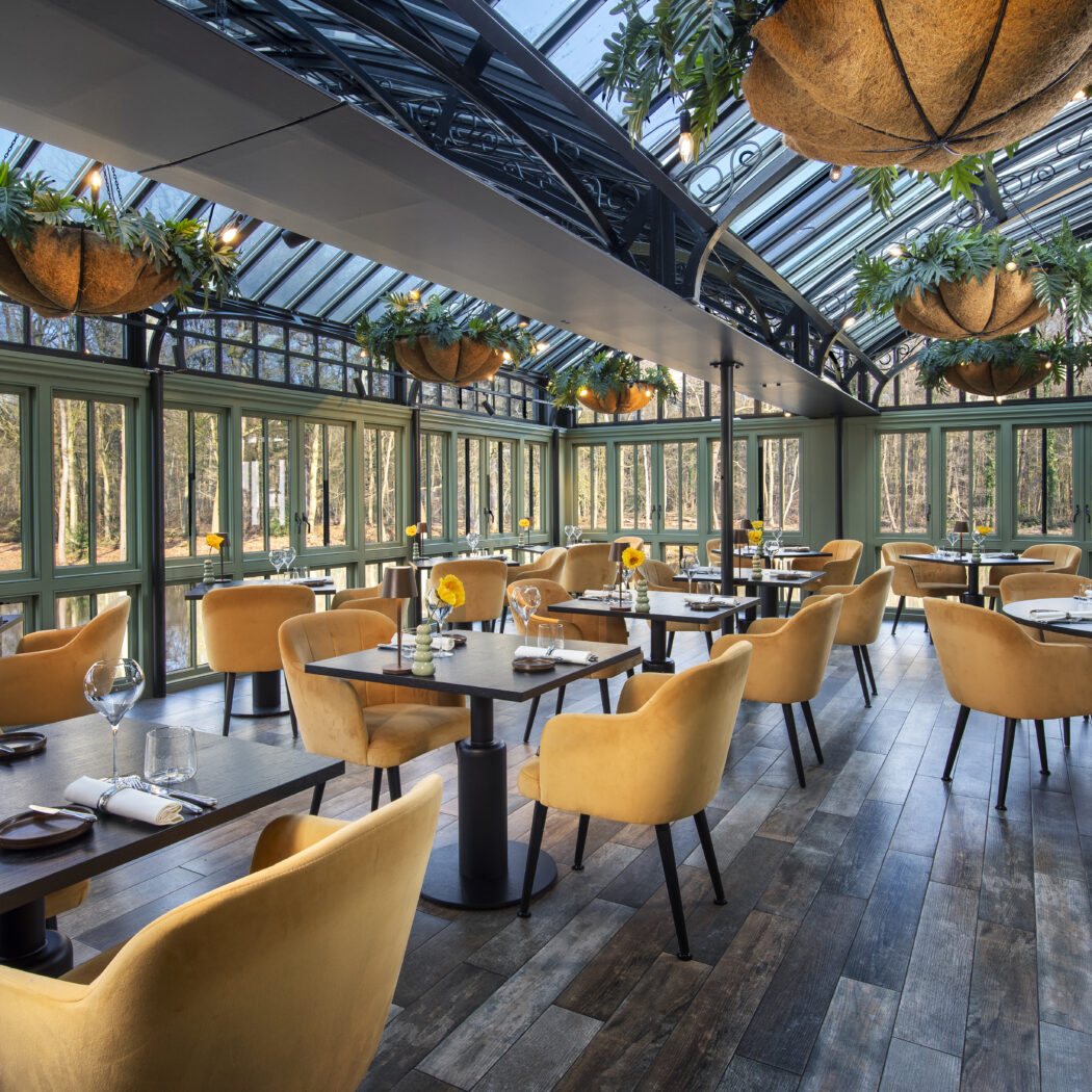Stijlvolle brasserie met okergele stoelen, glazen dak, hangplanten en panoramisch boszicht.