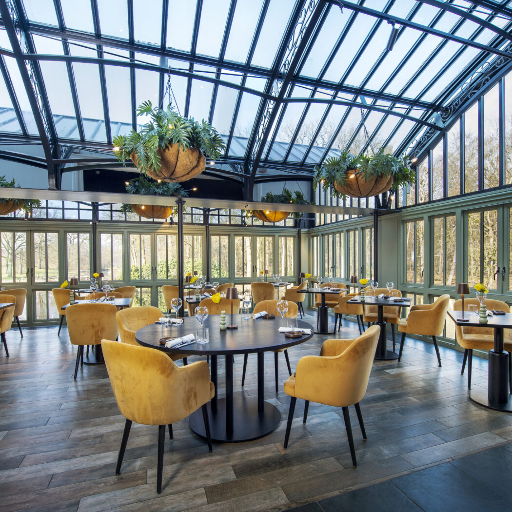 Ruime glazen serre restaurant met groene raamkozijnen en gele fluwelen stoelen aan gedekte tafels.