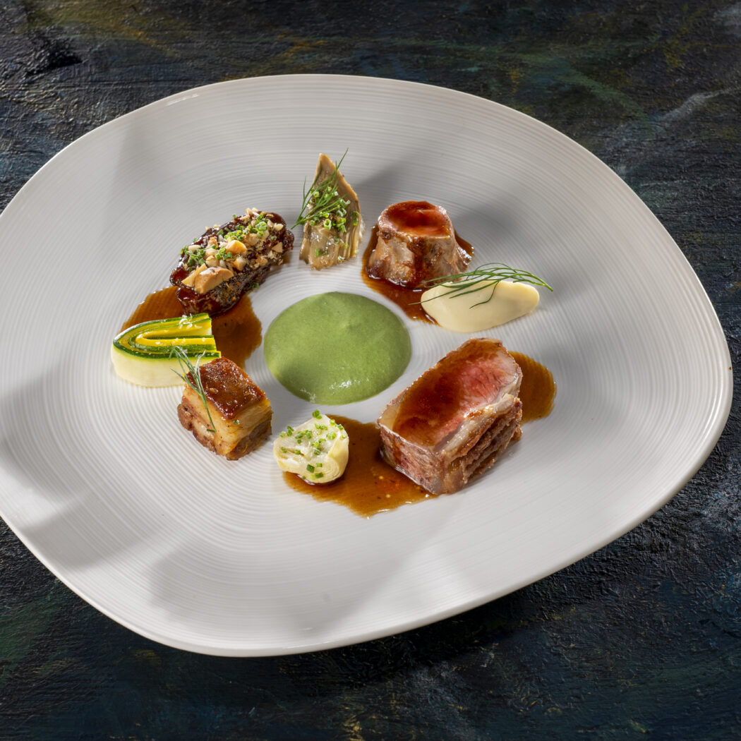 Elegant gepresenteerd gastronomisch vleesgerecht met diverse bereidingen, groene puree en garnituren op een wit bord.