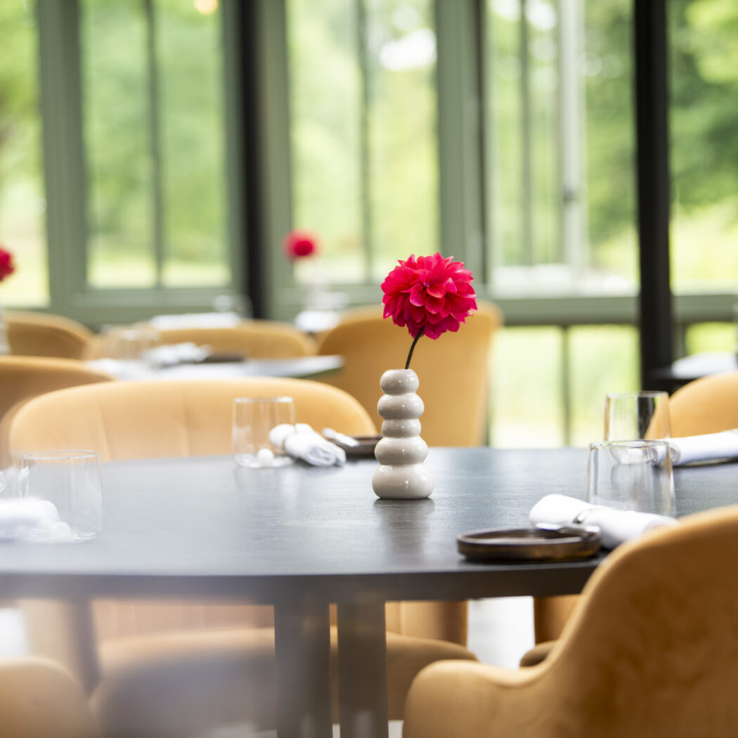 Gedekte restauranttafel met roze dahlia, glazen, okergele stoelen en groen uitzicht door grote ramen.