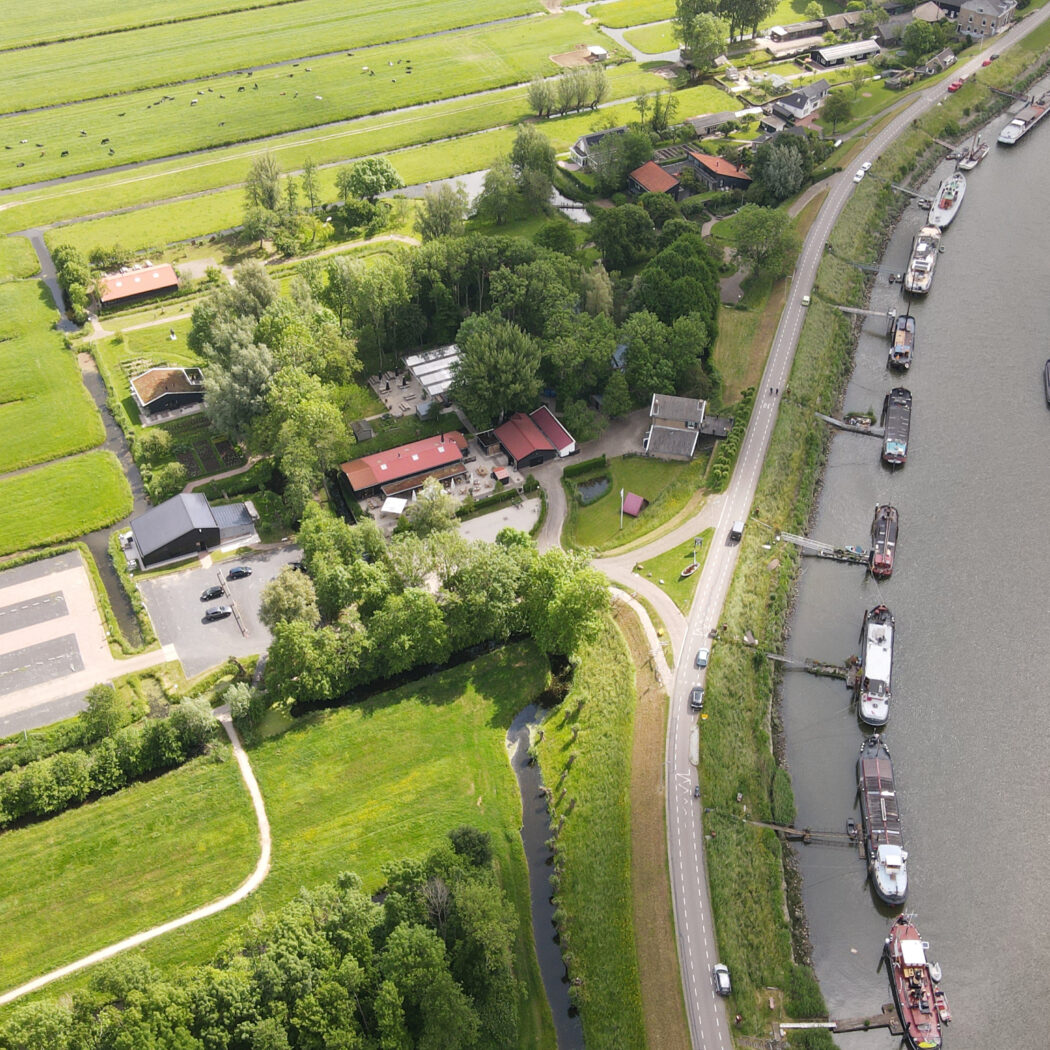 Luchtfoto van een rivier met tientallen schepen, een langsgelegen weg, groene landbouwgronden en bebouwing.