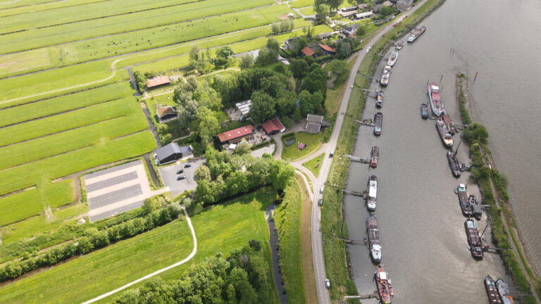 Luchtfoto van een rivier met tientallen schepen, een langsgelegen weg, groene landbouwgronden en bebouwing.