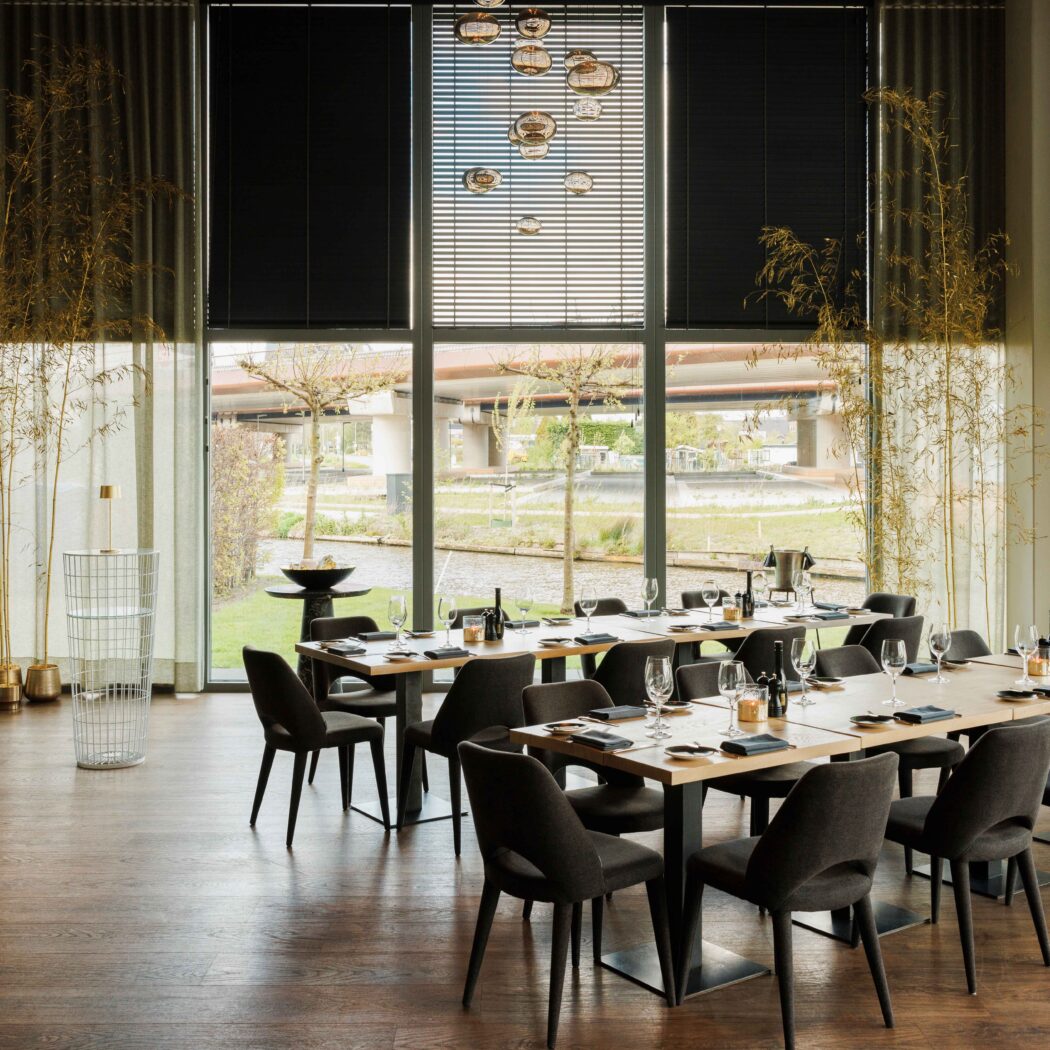Moderne eetzaal met lange gedekte houten tafels, donkere stoelen en grote ramen die uitkijken op een kanaal.
