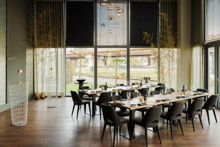 Moderne eetzaal met lange gedekte houten tafels, donkere stoelen en grote ramen die uitkijken op een kanaal.