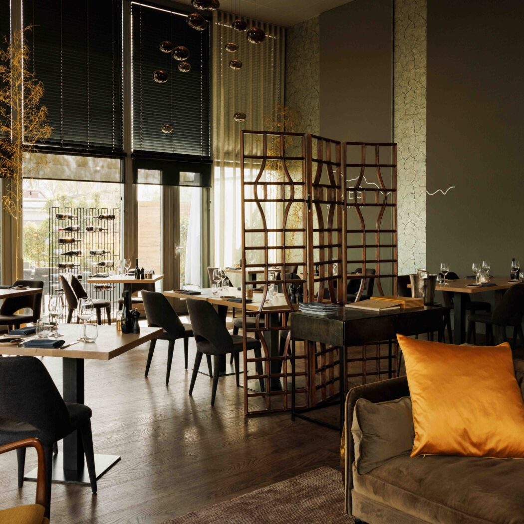 Sfeervol restaurant met lichte houten eettafels, donkere stoelen, grote ramen en een bank met oranje kussen.