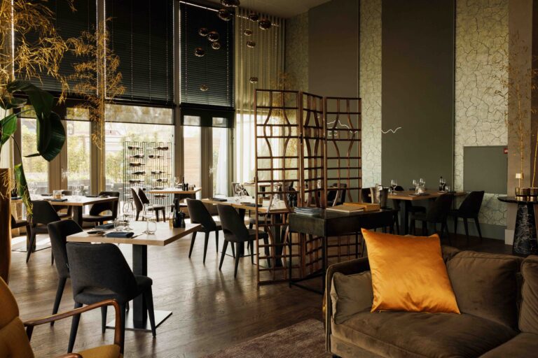 Sfeervol restaurant met lichte houten eettafels, donkere stoelen, grote ramen en een bank met oranje kussen.