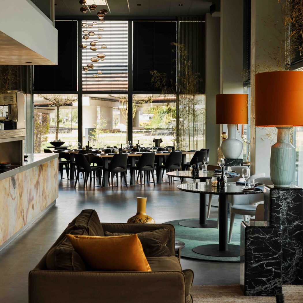 Modern restaurantinterieur met lange gedekte tafel, bruine bank, marmeren balie en opvallende oranje lampen.
