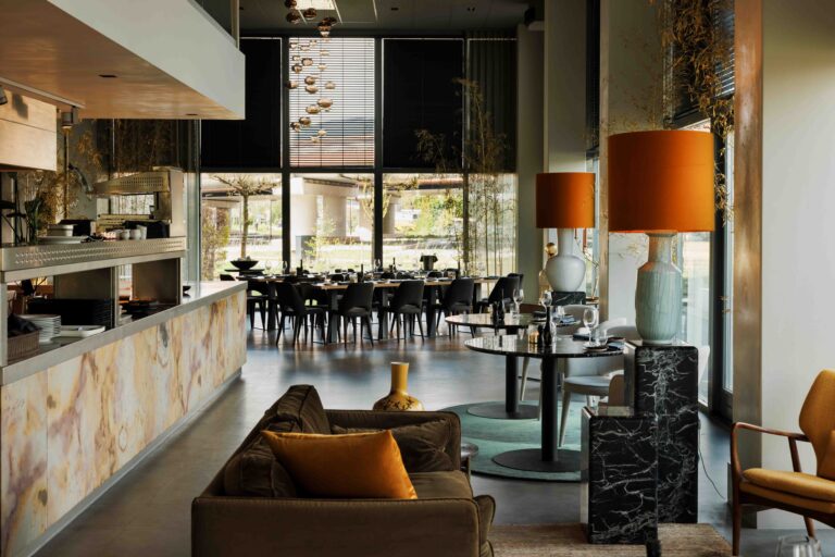 Modern restaurantinterieur met lange gedekte tafel, bruine bank, marmeren balie en opvallende oranje lampen.