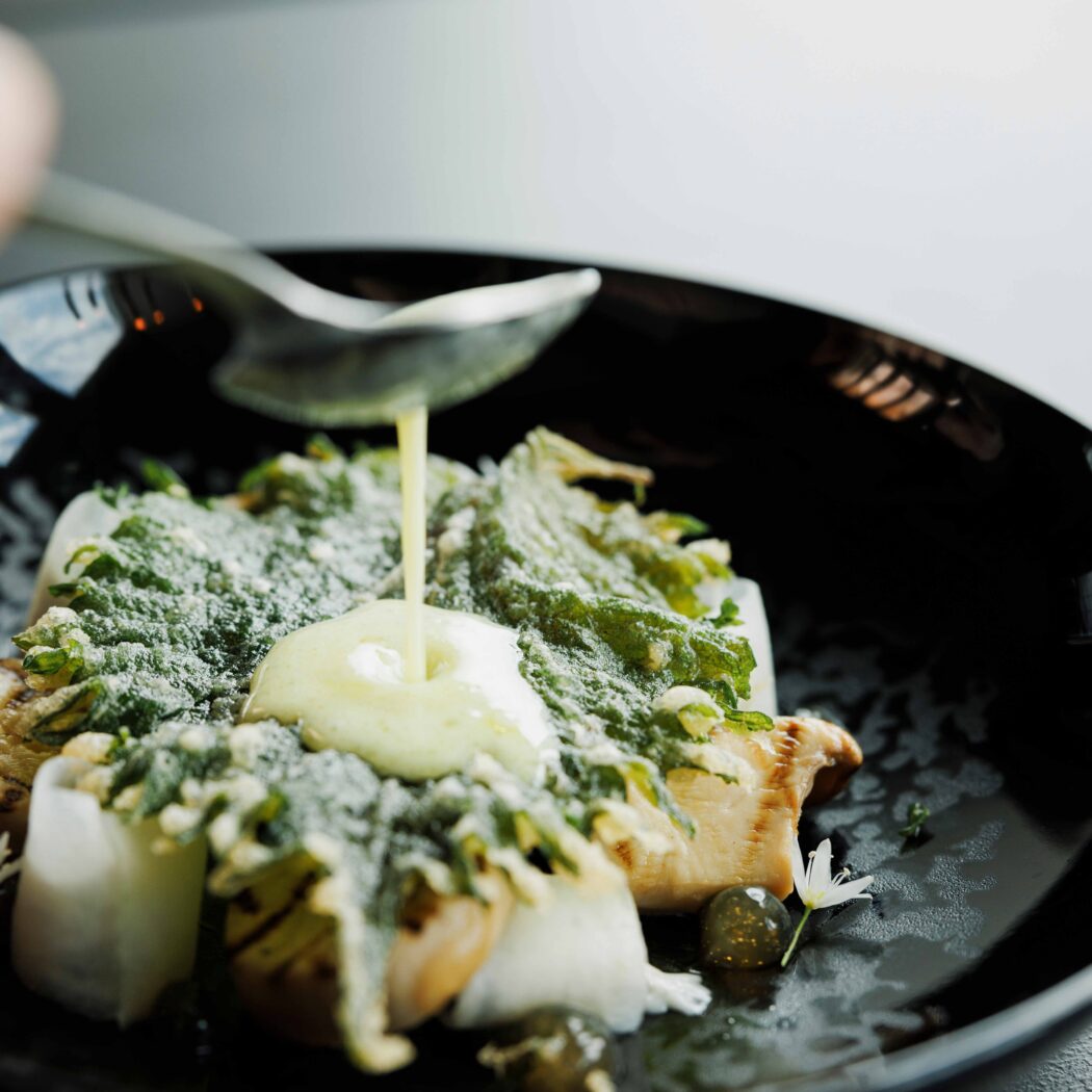 Een lepel giet romige saus over een culinair gerecht met gebakken paddenstoelen, knapperige groene kruiden en witte groenten op een zwarte schaal.
