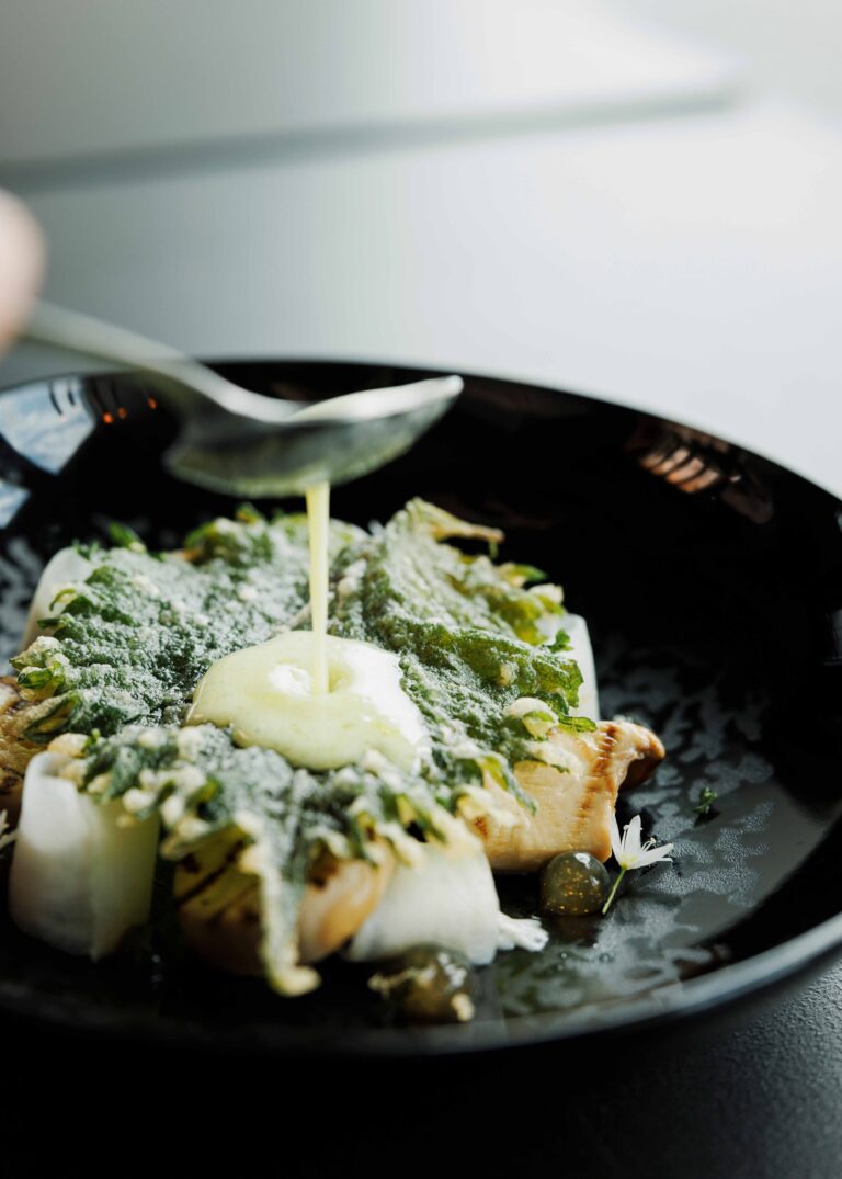 Een lepel giet romige saus over een culinair gerecht met gebakken paddenstoelen, knapperige groene kruiden en witte groenten op een zwarte schaal.