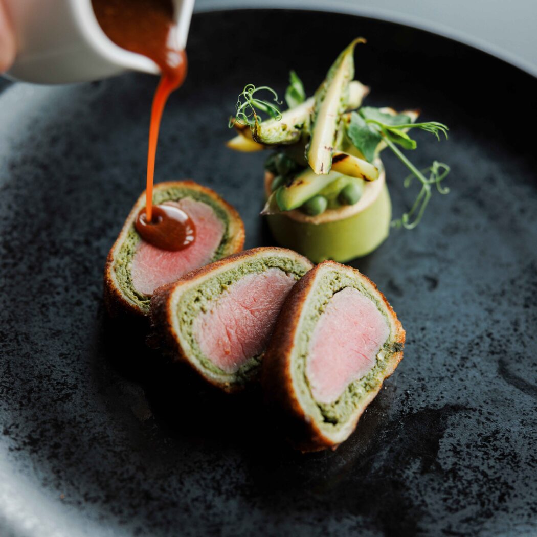 Malse plakjes vlees met groene vulling en krokante korst, geserveerd met gegrilde asperges; saus wordt eroverheen gegoten.