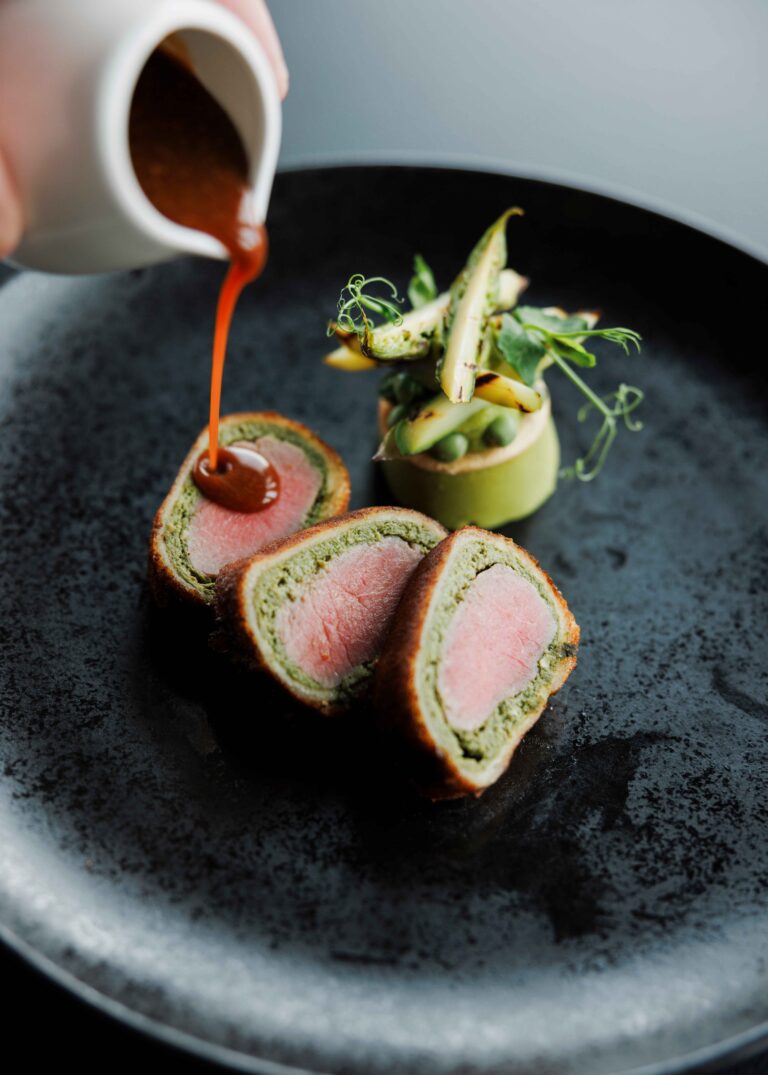 Malse plakjes vlees met groene vulling en krokante korst, geserveerd met gegrilde asperges; saus wordt eroverheen gegoten.