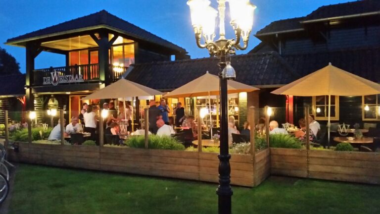 Restaurant De Wristaart met een sfeervol verlicht buitenterras en gasten onder beige parasols in de avond.