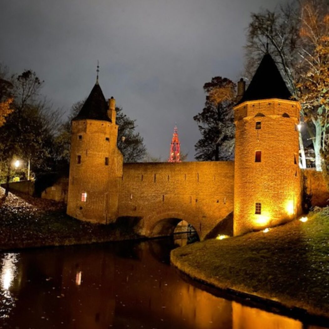 Historische bakstenen vesting met twee verlichte torens en een brug over een donkere gracht, rode spits in de verte.