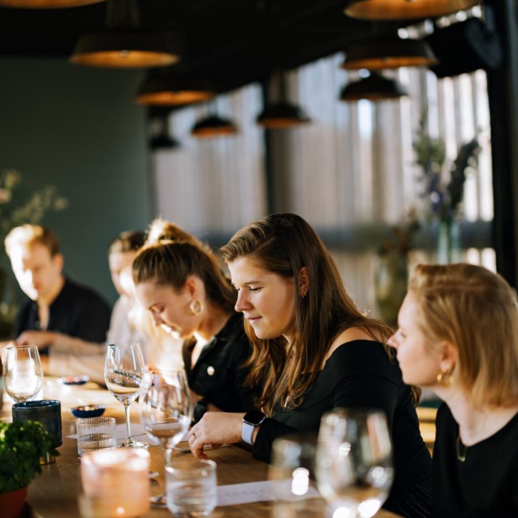 Jonge vrouwen geconcentreerd aan lange houten tafel met wijnglazen en kaarsen onder hanglampen.
