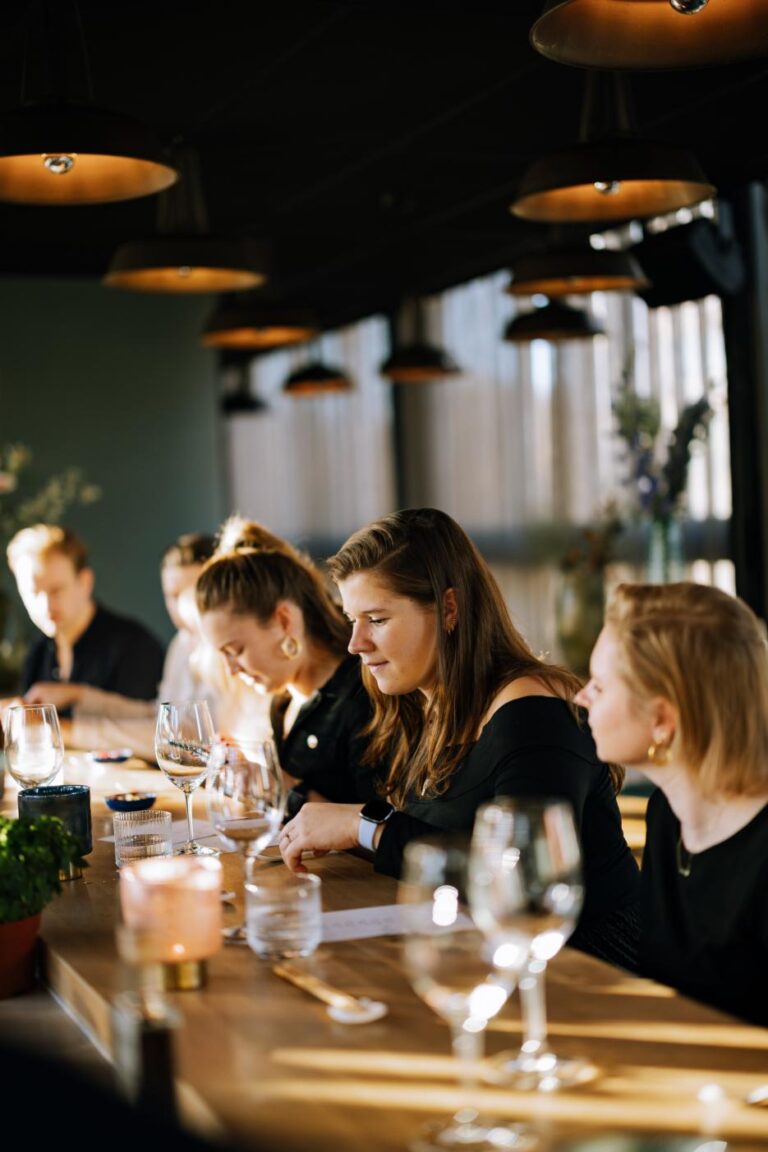 Jonge vrouwen geconcentreerd aan lange houten tafel met wijnglazen en kaarsen onder hanglampen.