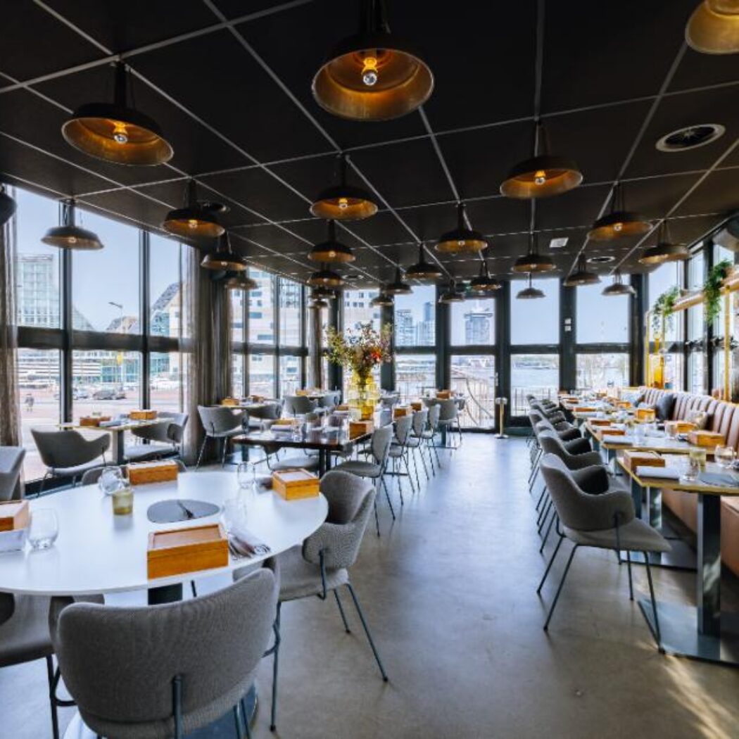 Modern restaurantinterieur met industriële hanglampen, bruine banken en panoramisch uitzicht op de stad en het water.