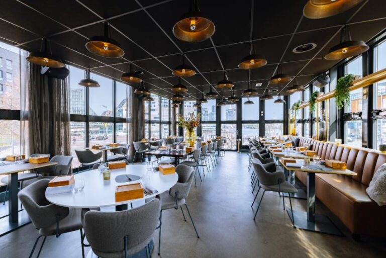 Modern restaurantinterieur met industriële hanglampen, bruine banken en panoramisch uitzicht op de stad en het water.