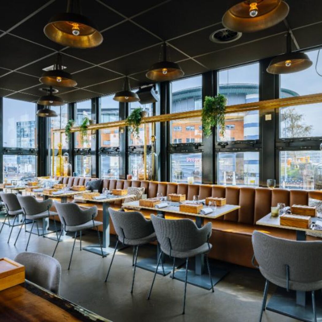 Modern restaurant interieur met bruine banken, grijze stoelen, gouden lampen en uitzicht door grote ramen.