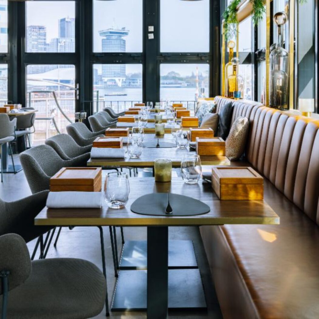 Stijlvol restaurant interieur met bruine banketbank, grijze stoelen en houten tafels; uitzicht op stad en water.