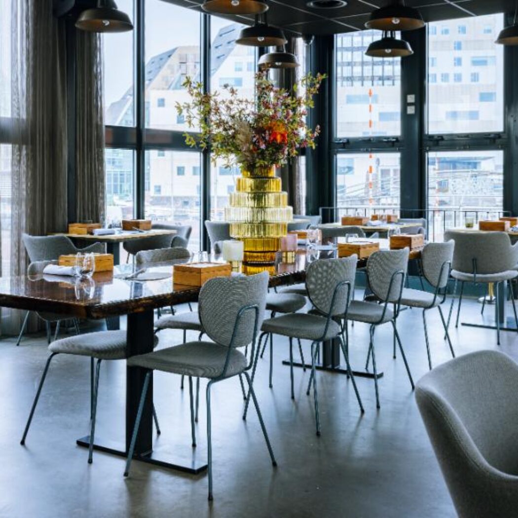 Modern restaurantinterieur met grote ramen, donkere tafels, grijze stoelen en een gouden vaas met bloemen.