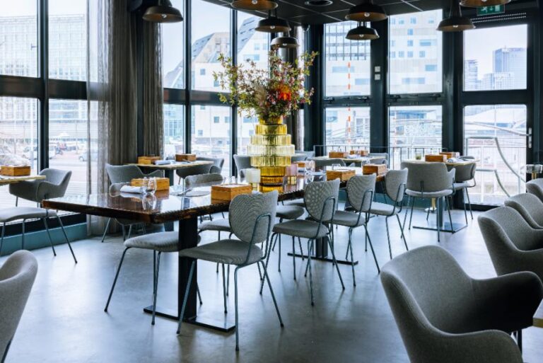 Modern restaurantinterieur met grote ramen, donkere tafels, grijze stoelen en een gouden vaas met bloemen.
