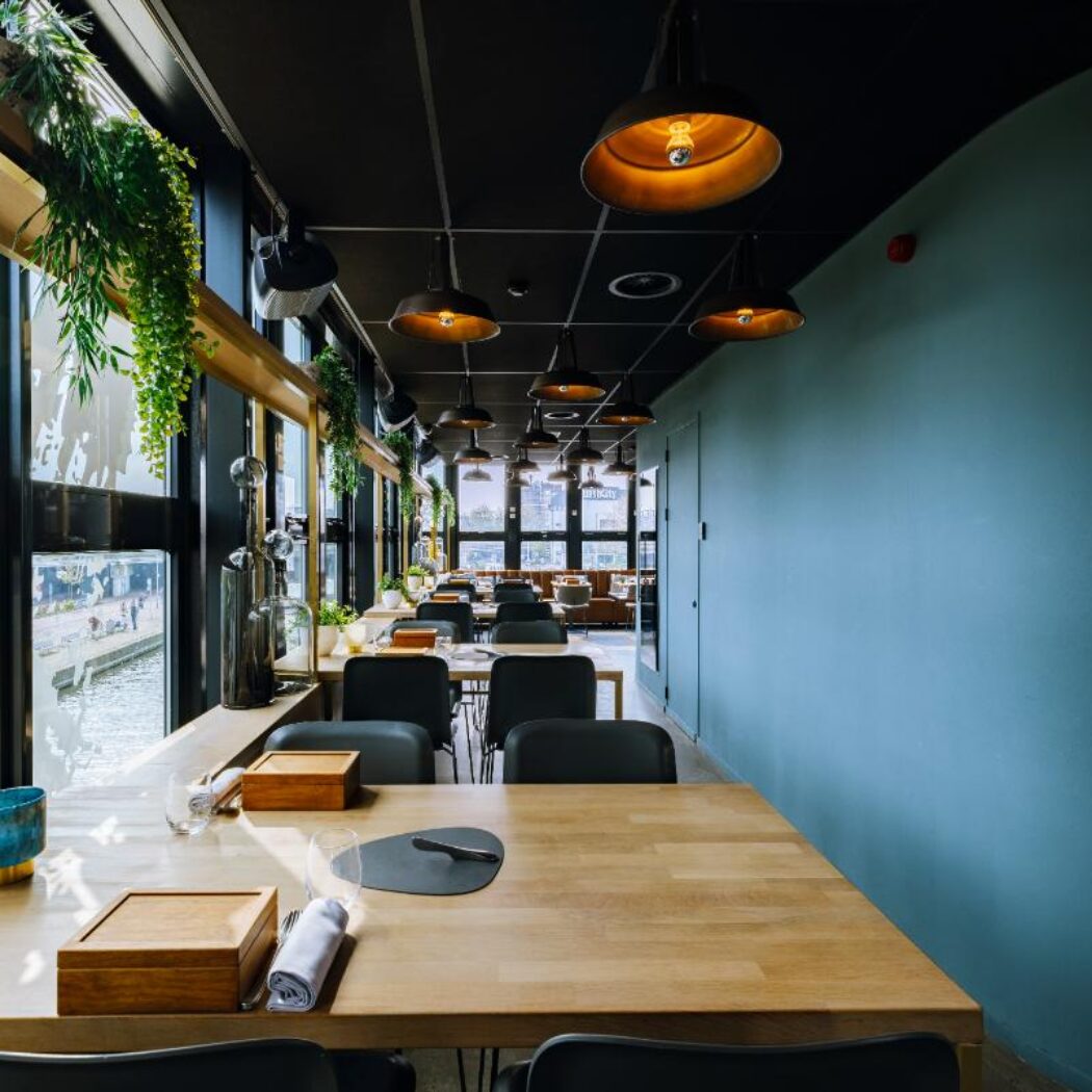 Lange, moderne restaurantruimte met houten tafels, zwarte stoelen, industriële lampen en uitzicht op water.