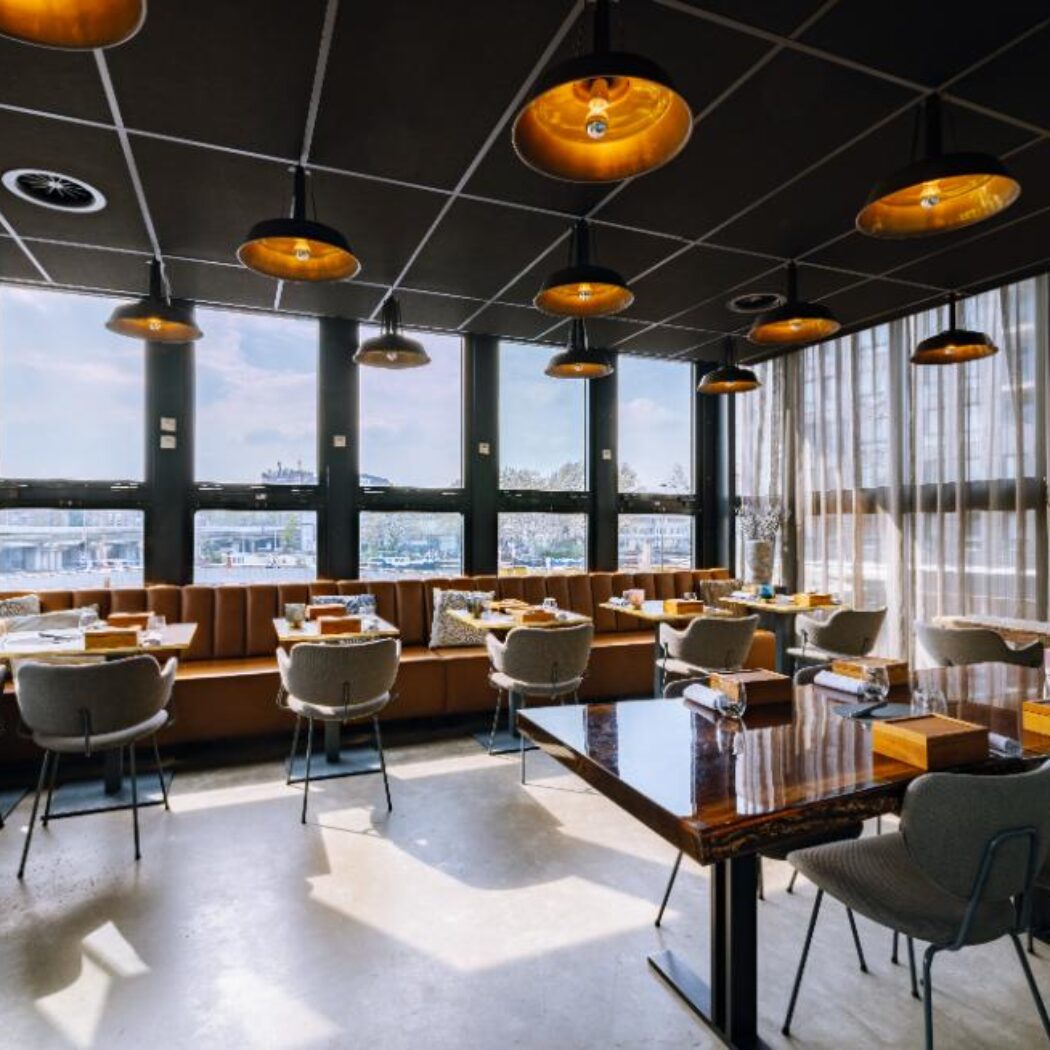 Licht restaurant met industriële hanglampen met gouden binnenkant, bruine banken en houten tafels bij grote ramen.