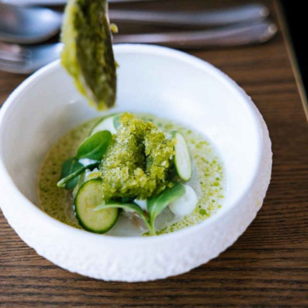 Een lepel voegt groene granita toe aan een amuse van courgette en kruiden in een witte kom op een houten tafel.