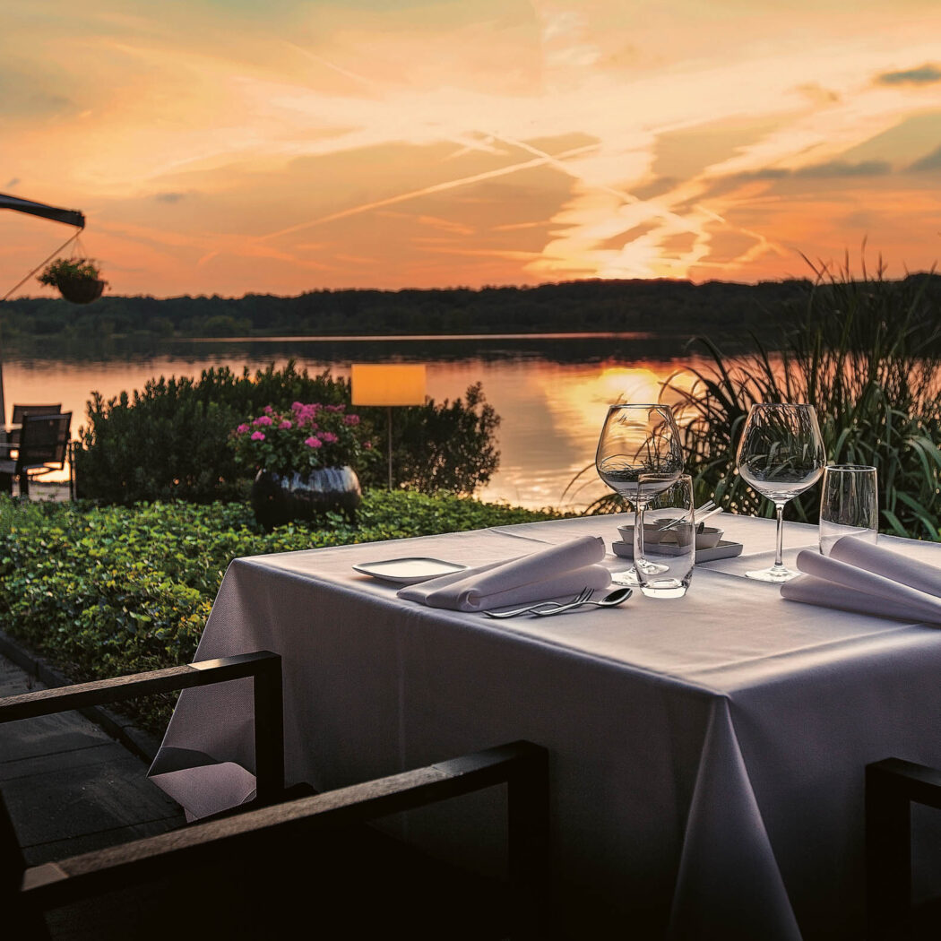 Gedekte tafel met wijnglazen op buitenterras met oranje zonsondergang boven een meer.