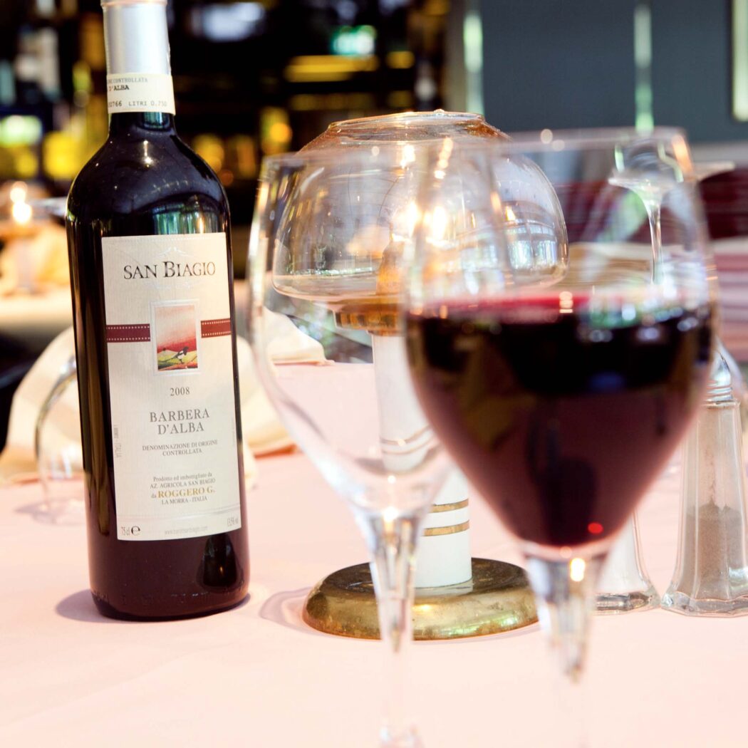 Fles rode wijn San Biagio Barbera dAlba 2008 en twee wijnglazen, één gevuld, op een roze tafelkleed.