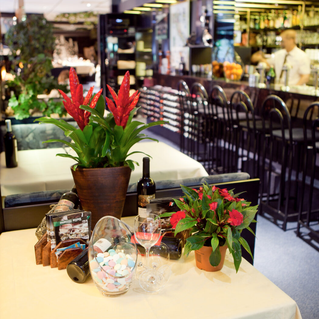 Interieur van een restaurant met tafeldecoratie: rode bloemen, wijn, snoepjes, en een bar met barkrukken en een barman.