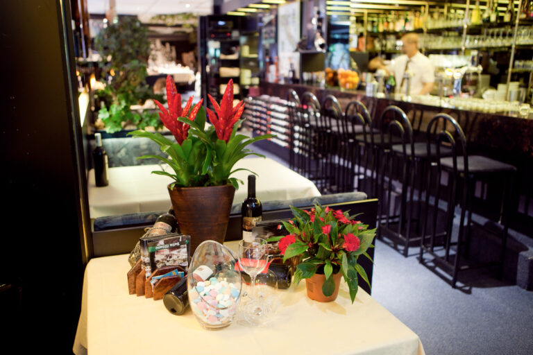 Interieur van een restaurant met tafeldecoratie: rode bloemen, wijn, snoepjes, en een bar met barkrukken en een barman.