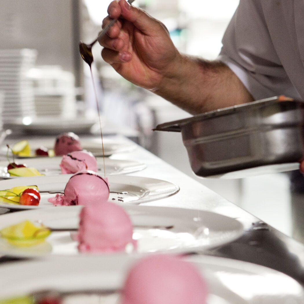 Chef dresseert roze ijsbollen met donkere saus en fruit op meerdere borden in een professionele keuken.