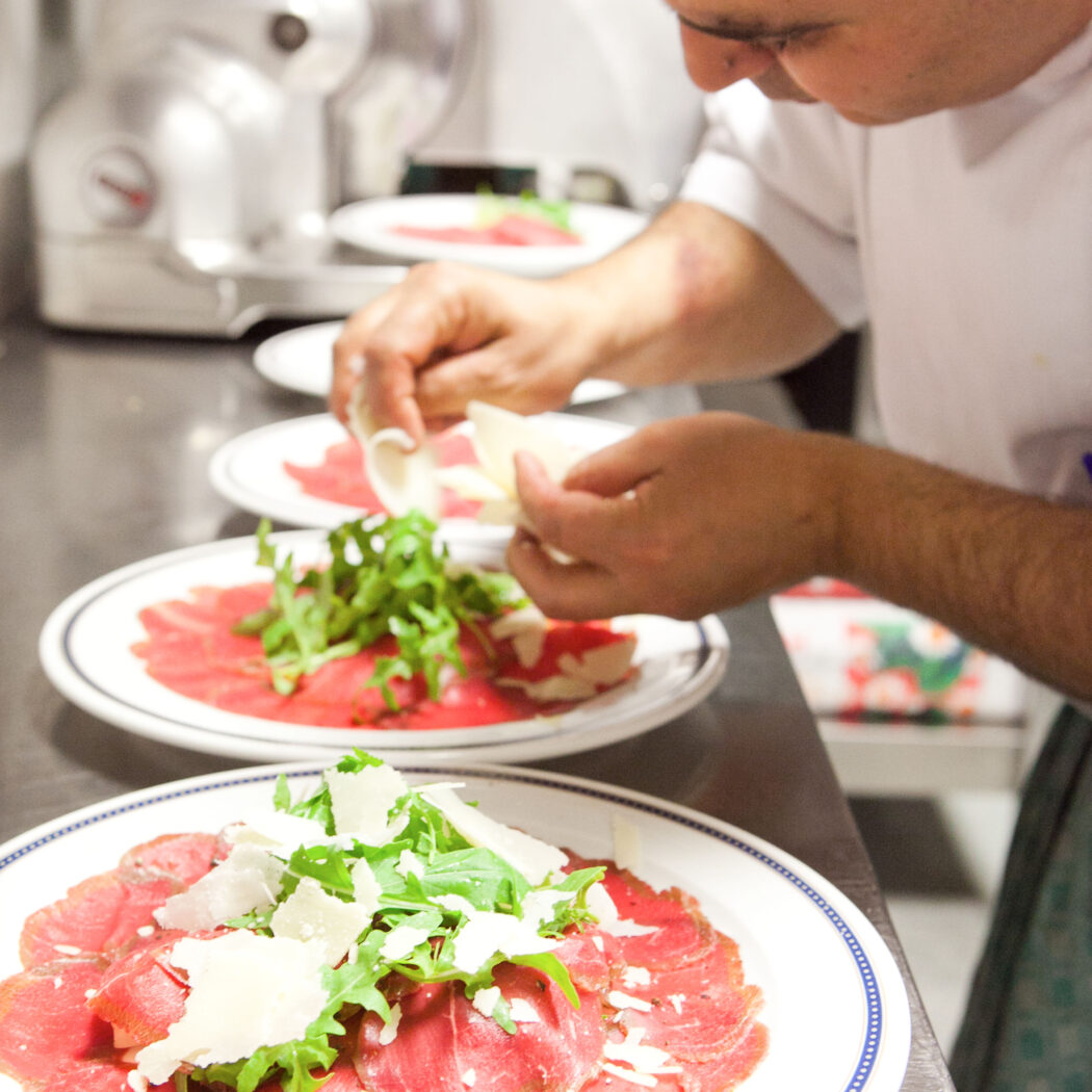 Kok bereidt meerdere borden met carpaccio, rucola en geschaafde Parmezaanse kaas.