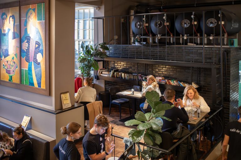 Gezellig café-interieur met etende en drinkende gasten, biertanks op een entresol en een kleurrijk schilderij.