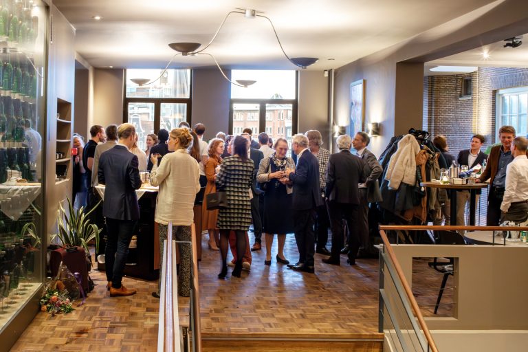 Een groep mensen netwerkt en socialiseert in een lichte, moderne evenementenruimte met grote ramen en houten vloer.