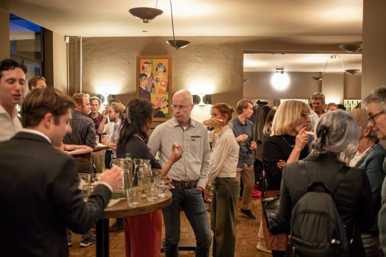 Gasten praten en drinken bij een ronde tafel met kannen en glazen, onder een kleurrijk schilderij met MIND.