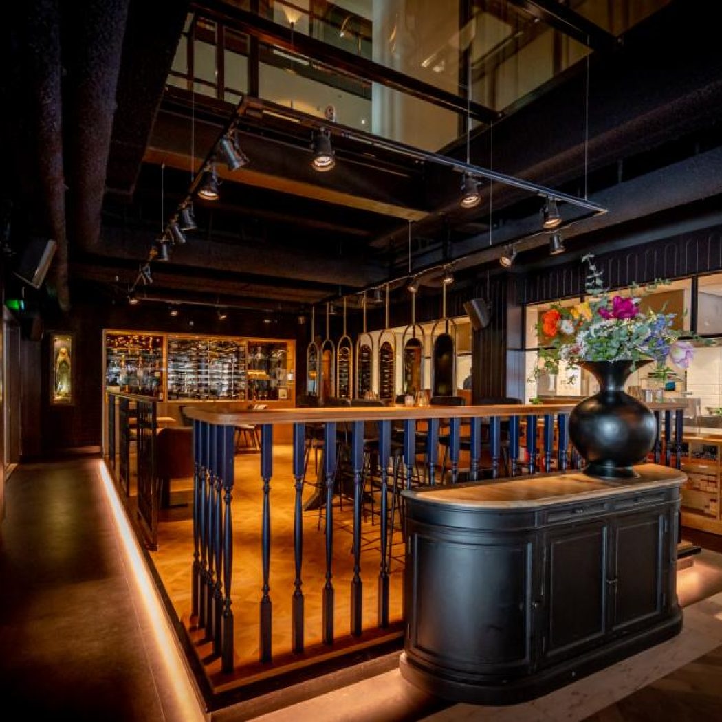 Modern restaurant met donkere houten bar, open keuken, grote wijnkast en kleurrijke bloemen op een balie.
