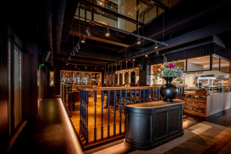 Modern restaurant met donkere houten bar, open keuken, grote wijnkast en kleurrijke bloemen op een balie.