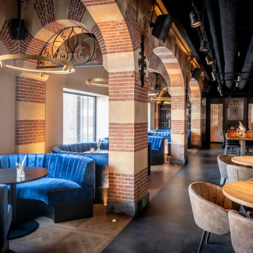 Modern restaurantinterieur SAINT met blauwe fluwelen banquettes, bakstenen bogen en warme verlichting.