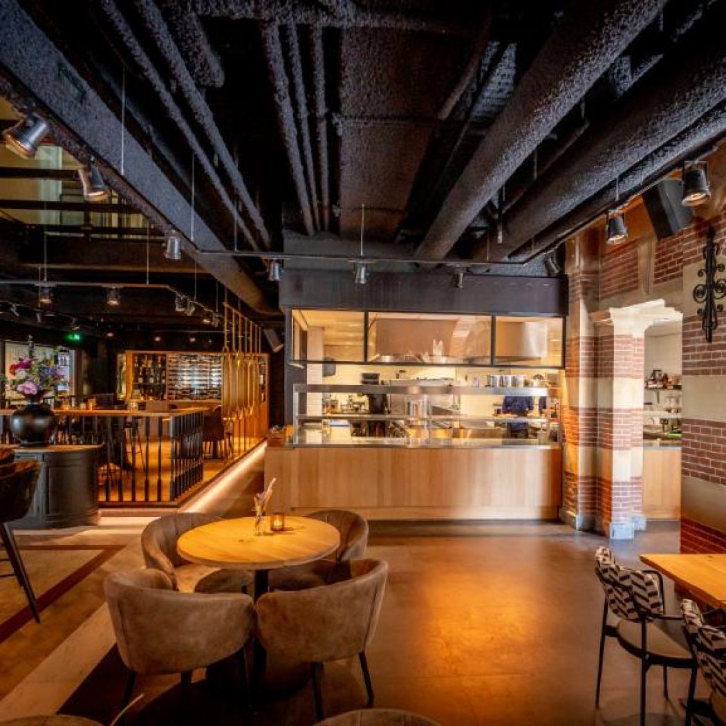 Modern restaurant interieur met een open keuken, bakstenen muren, houten tafels en zichtbare leidingen aan het zwarte plafond.