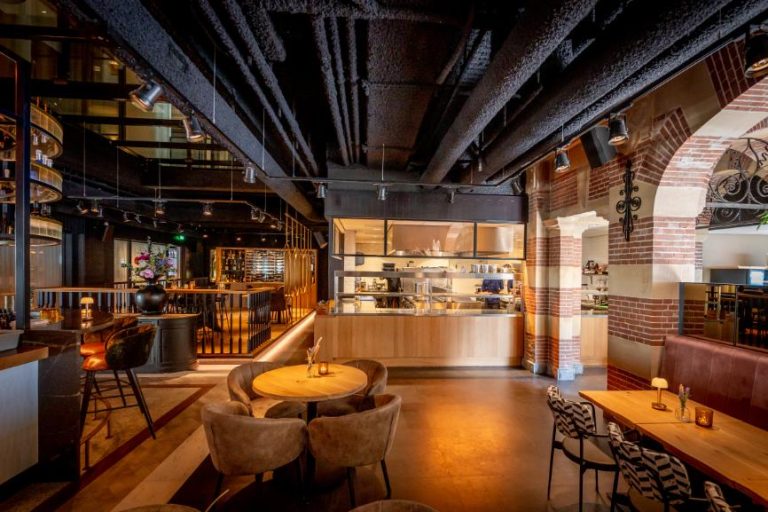 Modern restaurant interieur met een open keuken, bakstenen muren, houten tafels en zichtbare leidingen aan het zwarte plafond.