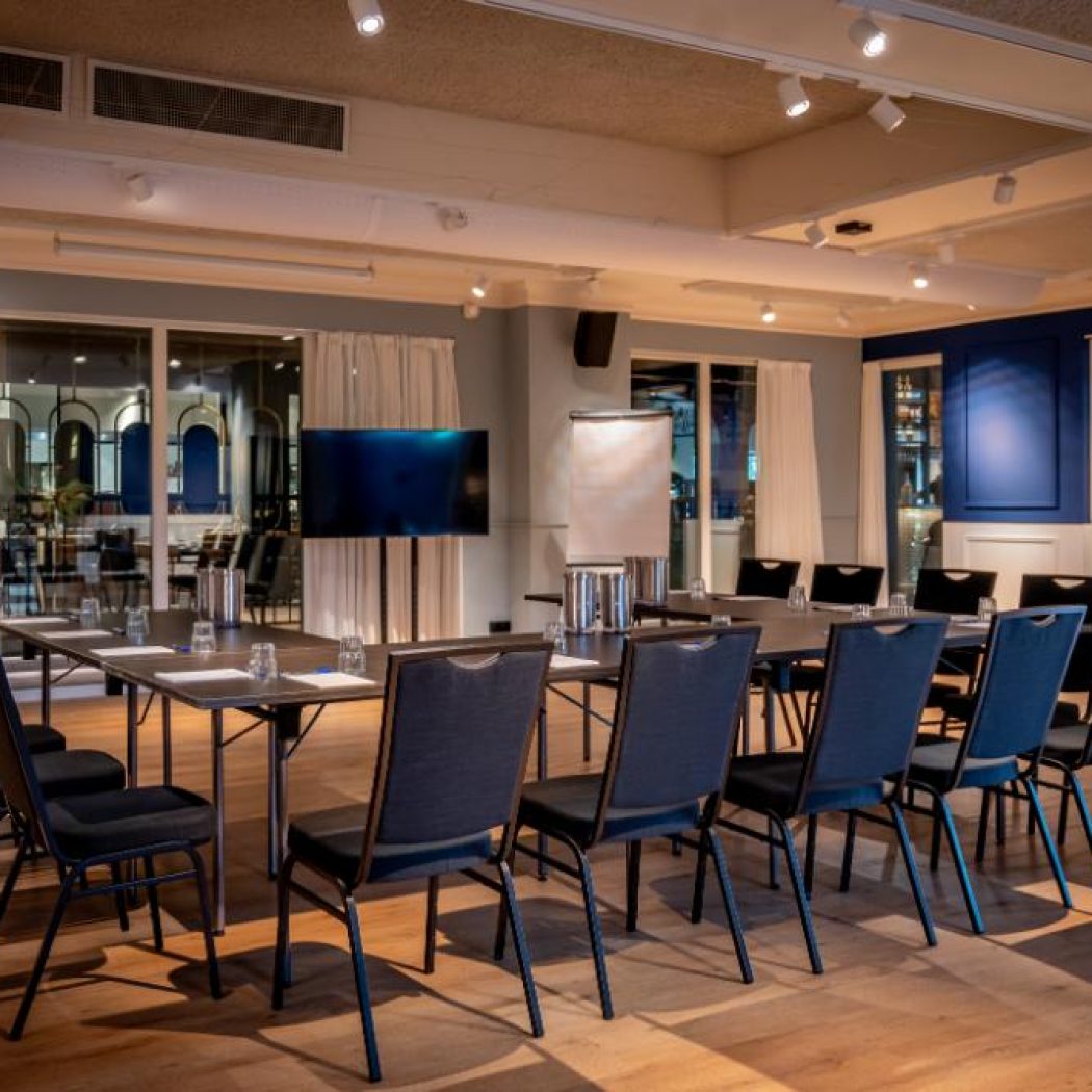 Moderne conferentiezaal met U-vormige tafelopstelling, donkere stoelen, een tv-scherm en flip-over op een houten vloer.