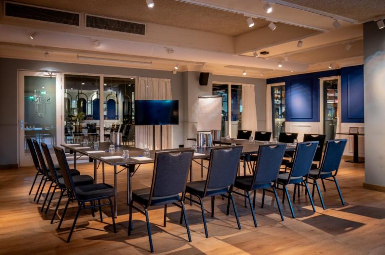 Moderne conferentiezaal met U-vormige tafelopstelling, donkere stoelen, een tv-scherm en flip-over op een houten vloer.