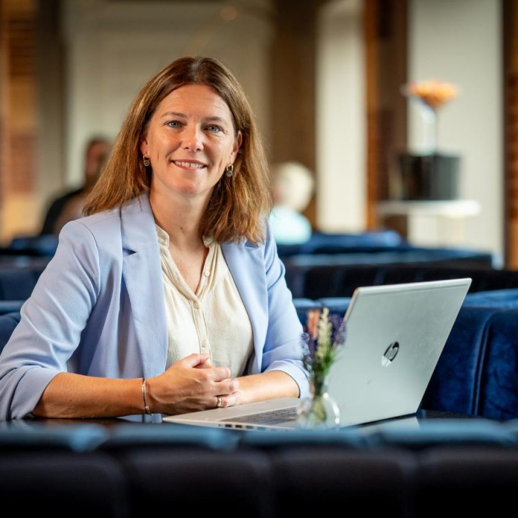 Glimlachende vrouw in lichtblauwe blazer zit aan tafel met HP laptop in een sfeervolle lounge.
